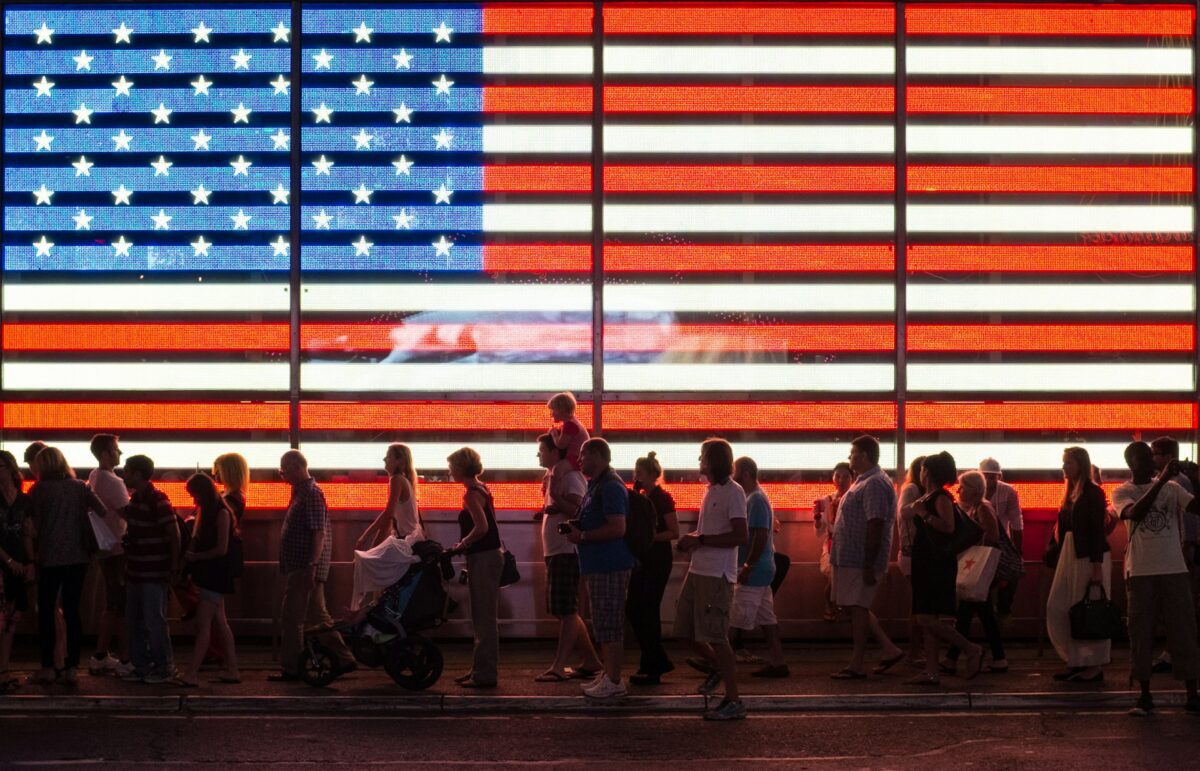People walking beneath a large illuminated American flag at night.