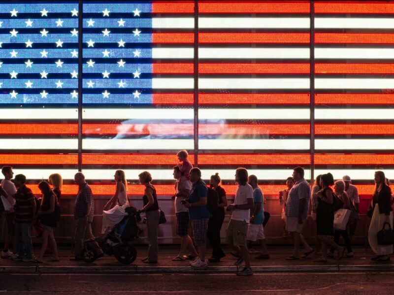 People walking beneath a large illuminated American flag at night.